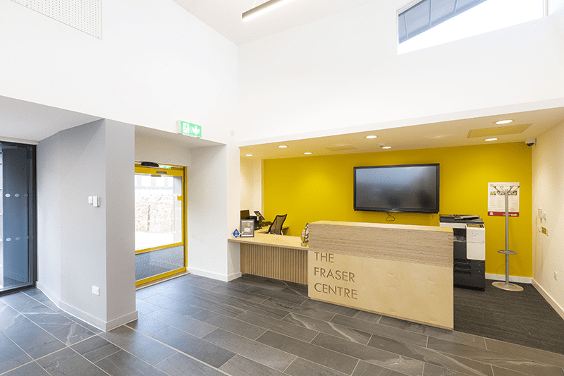 Reception desk at the Fraser Centre with a bright yellow wall behind Reception desk at the Fraser Centre with a bright yellow wall behind